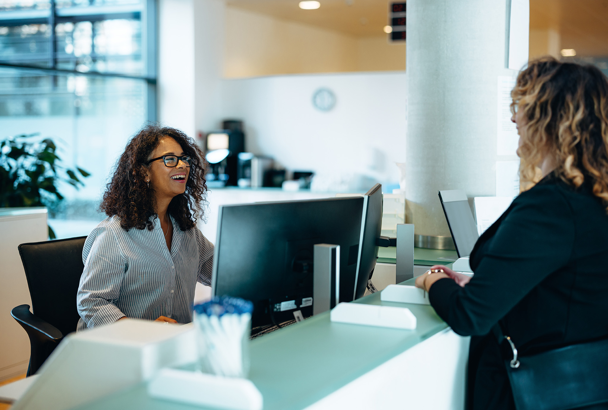 A receptionist sitting behind a desk is smiling and speaking with a woman standing on the opposite side. Both are indoors in a well-lit office environment.