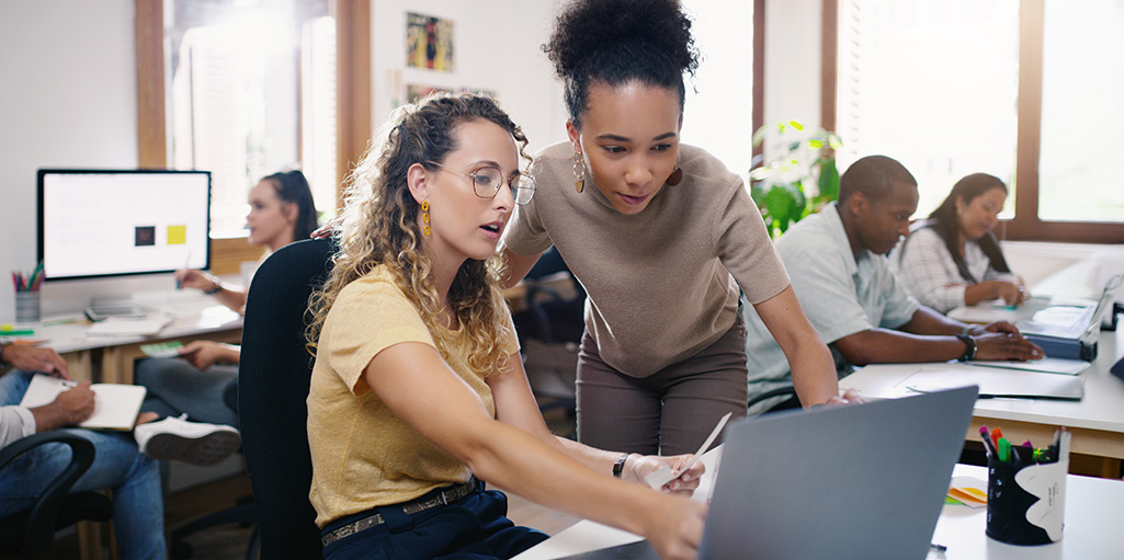 Student and trainer working together at a desk with a laptop, while three other people work in the background in a busy office setting.